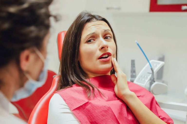 Woman in dental chair holding cheek with concerned expression during dental visit